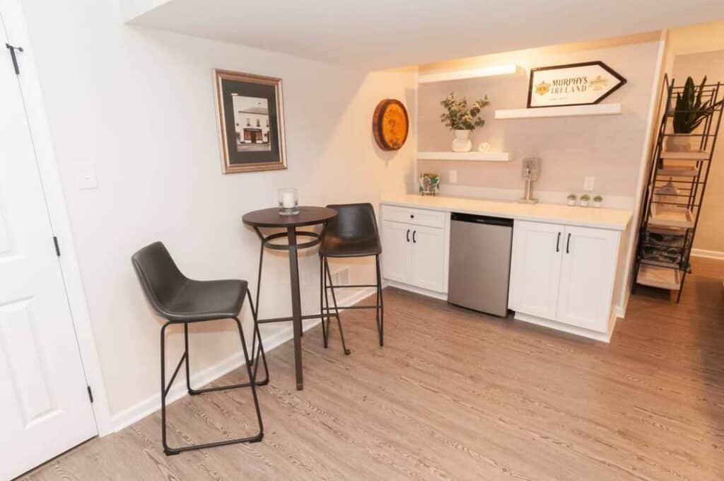 a small kitchen area with white cabinets, a mini fridge, a countertop with decor, a round table with two modern chairs, wall art, and a wooden floor. a staircase is visible in the background.