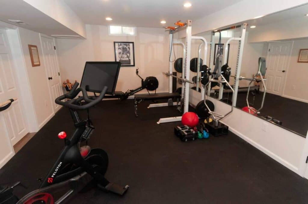 a home gym with a peloton bike, rowing machine, weightlifting rack, various free weights, a red exercise ball, kettlebells, and a large wall mirror. the room has white walls and closed double doors.