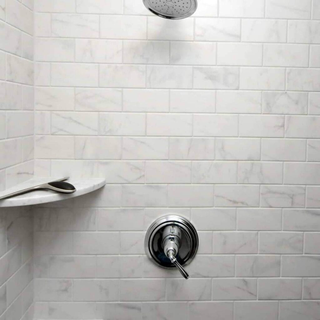 a tiled shower with a round showerhead at the top, a metal temperature control handle below, and two spoons resting on a small corner shelf.