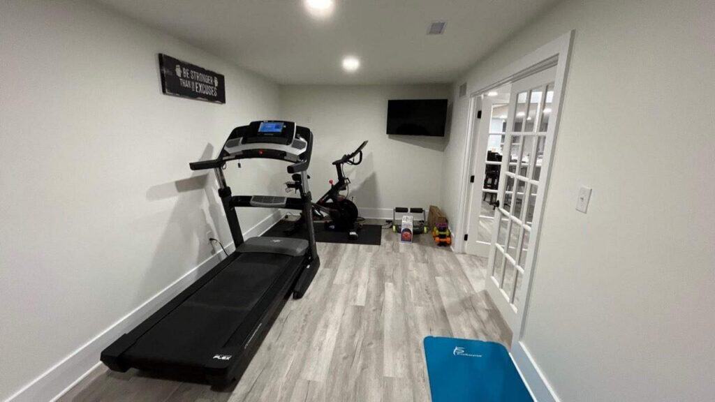 a home gym with a treadmill, exercise bike, wall mounted tv, free weights, yoga mat, and light wood flooring in a bright room with white walls and glass double doors.