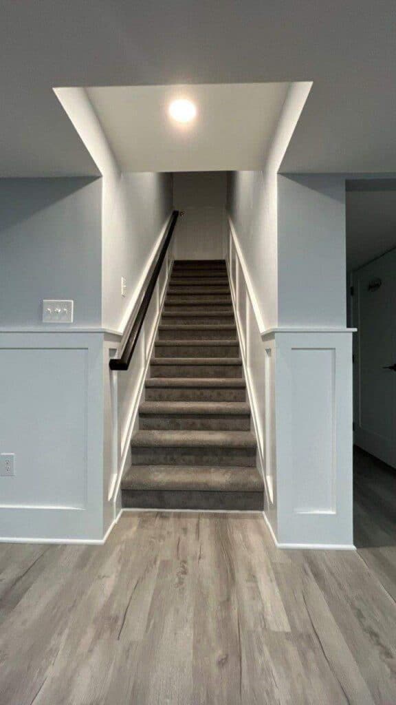 a staircase with gray carpet and white walls leads upward from a basement with light gray wood style flooring and white paneling. a single ceiling light illuminates the stairs.