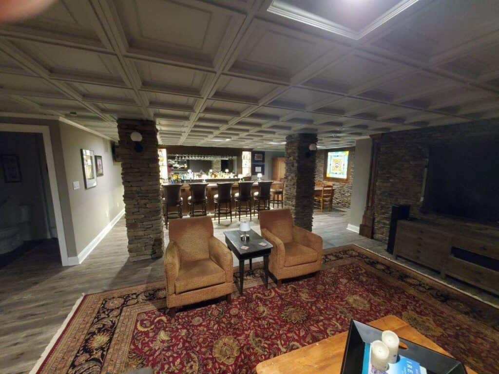 a cozy basement with a bar area featuring high stools, two armchairs with a small table, stone pillars, wood flooring, a large rug, and a tv stand. the ceiling has a grid pattern with recessed lighting.