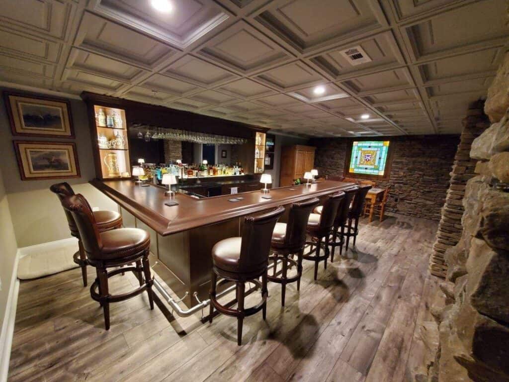 a cozy home bar with a u shaped wooden counter, high chairs, shelves stocked with bottles and glasses, and a stained glass window on a stone wall. wooden floors and framed art add to the warm atmosphere.