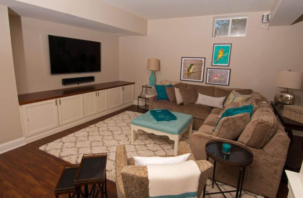 a cozy living room with a beige sectional sofa, accent pillows, a patterned rug, a turquoise ottoman, wall art with birds, a flat screen tv, and lamps on side tables, all in a neutral and teal color scheme.