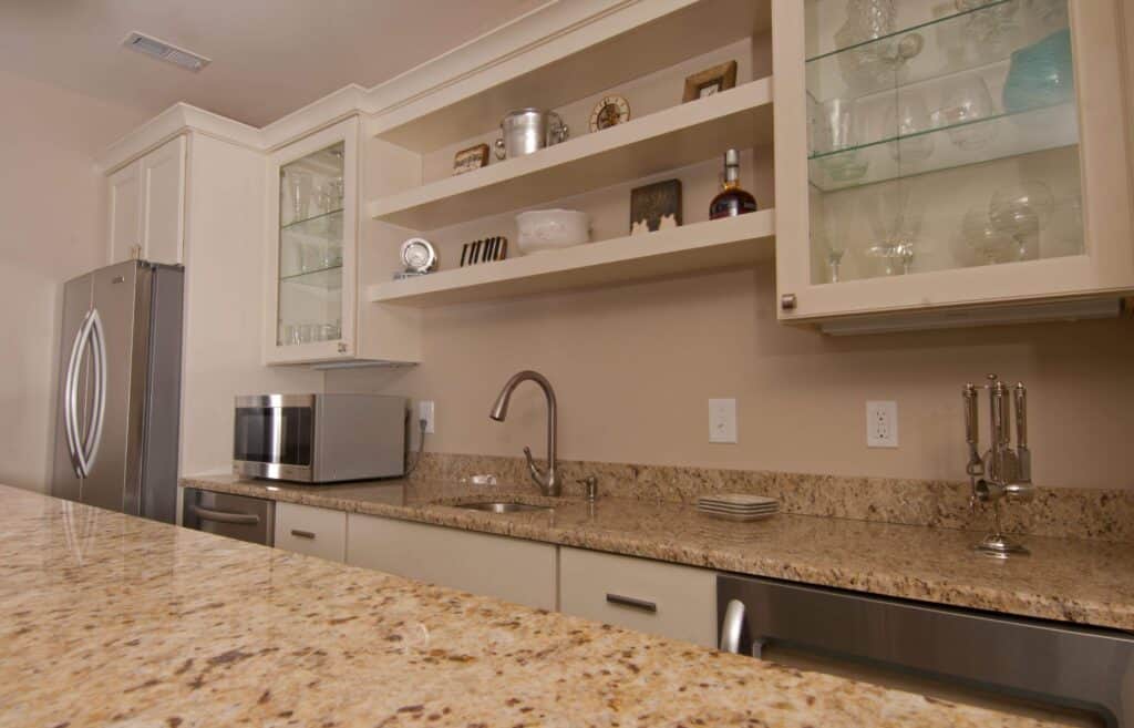 modern kitchen with beige granite countertops, stainless steel appliances, a microwave, sink, and open shelves displaying glassware, dishes, and decorative items. cabinets have glass doors, and the area looks clean and organized.