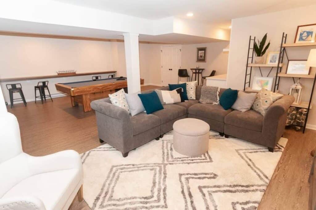 a modern, cozy basement living area with a gray sectional sofa, blue and patterned pillows, a round ottoman on a geometric rug, a white armchair, wall art, a pool table, and bar stools at a counter in the background.