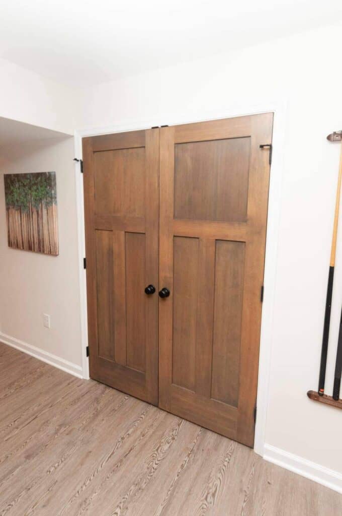 a pair of closed, wooden double doors with dark knobs, set in a white frame against a light colored wall. the floor is light wood, and a partial painting and pool cues are visible on the wall.