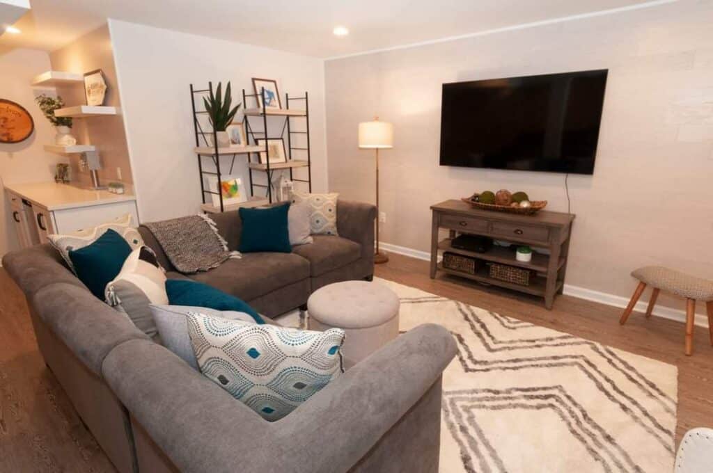 a cozy living room with a gray sectional sofa, patterned cushions, a round ottoman, a geometric rug, a wall mounted tv, a wooden tv stand, a floor lamp, and a decorative shelving unit with plants and books.