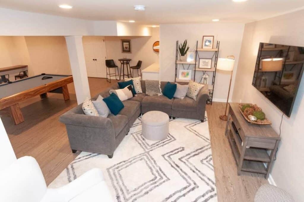 a cozy basement living area with a gray sectional sofa, blue and patterned pillows, a round ottoman, a patterned rug, a tv mounted on the wall, a wooden console, and a pool table in the background.