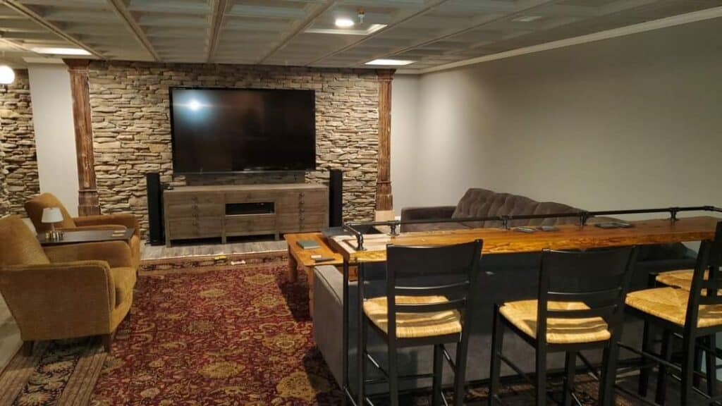 a cozy home theater with a large tv on a stone accent wall, surrounded by a brown sofa, tan armchair, a wooden console, and a bar with high chairs. the space features warm lighting and a patterned rug.