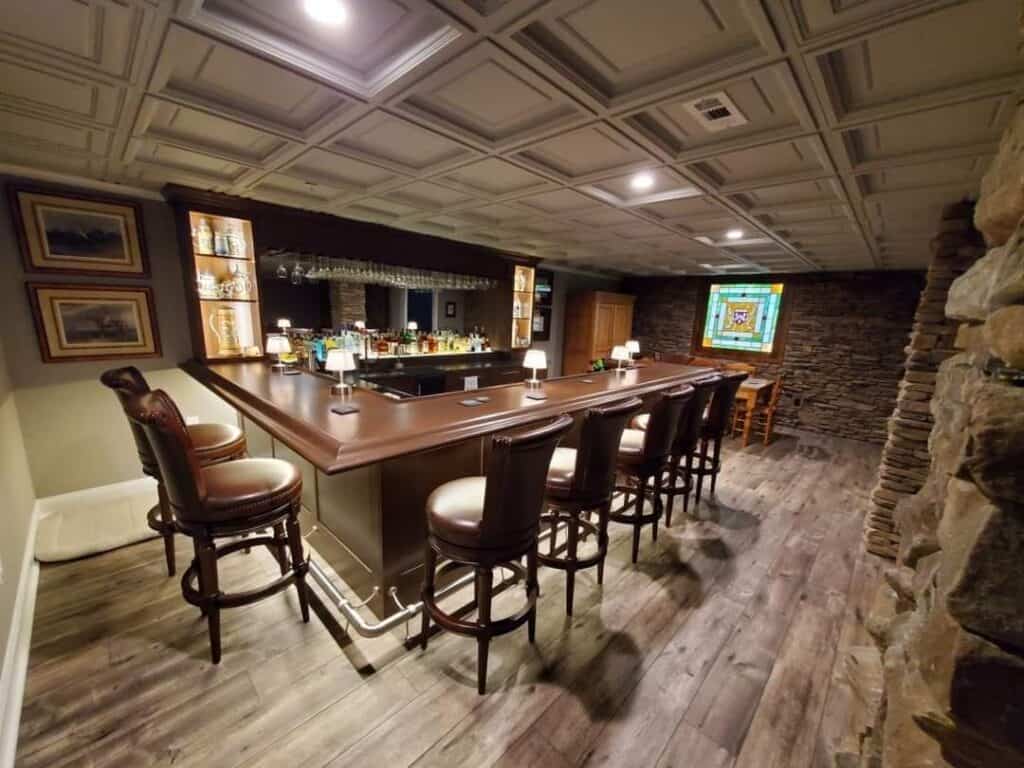 a home bar with wooden stools, a large counter, and shelves with liquor bottles and glasses. the room has wood floors, stone walls, framed art, and a stained glass window above a small table and chairs in the corner.