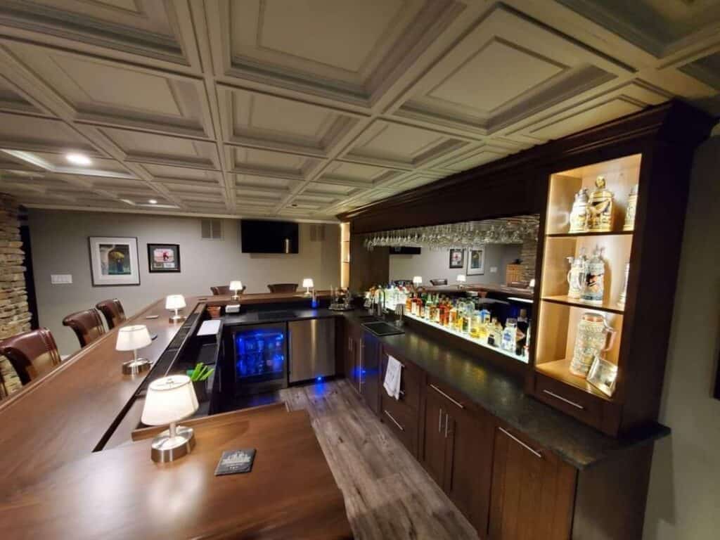 a home bar with wooden counters, bar stools, illuminated shelves of liquor bottles, glassware hanging above, small table lamps on the counter, and framed pictures on the wall, creating a cozy atmosphere.