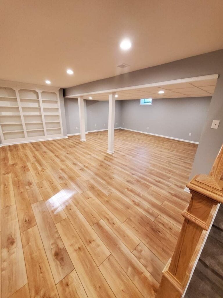 spacious finished basement with light wood flooring, gray walls, built in white shelves, recessed lighting, two white support columns, and a section of stairs with a wooden banister in the foreground.