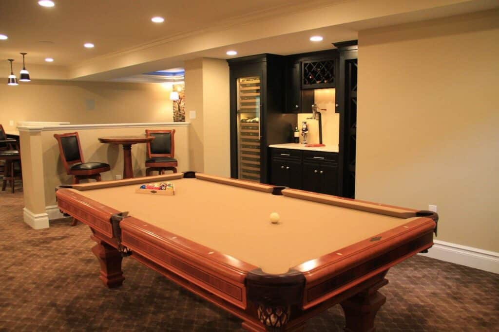 a basement game room with a tan felt pool table in the center, a small bar area with wine storage and sink in the back, and two chairs with a high table along a half wall. warm lighting and beige walls create a cozy atmosphere.