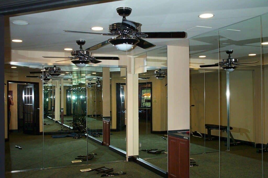 a room with mirrored walls reflecting ceiling fans, columns, and exercise equipment on the carpeted floor. the mirrors create multiple reflections, giving the space a larger, more complex appearance.
