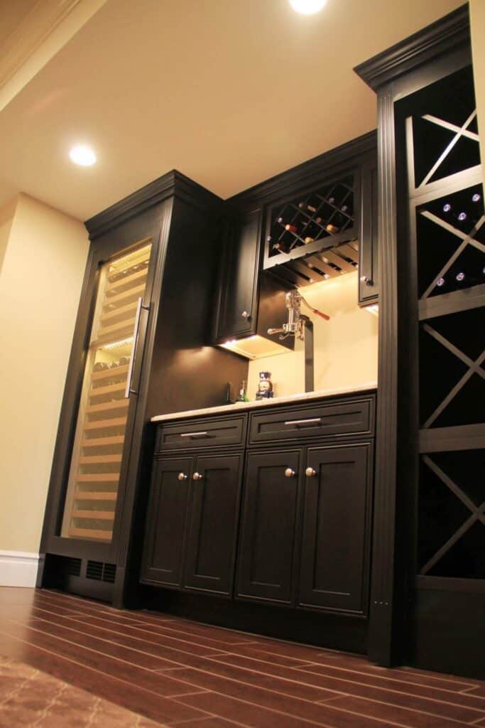 a built in home wine bar features dark cabinets, a tall wine fridge with glass door, a small countertop, and wine racks above and beside the counter, all set against beige walls and brown tile flooring.