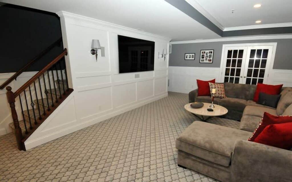 a cozy living room with a gray sectional sofa accented by red pillows, a round coffee table, a wall mounted tv, white paneled walls, patterned carpet, and a wooden staircase with black railing.
