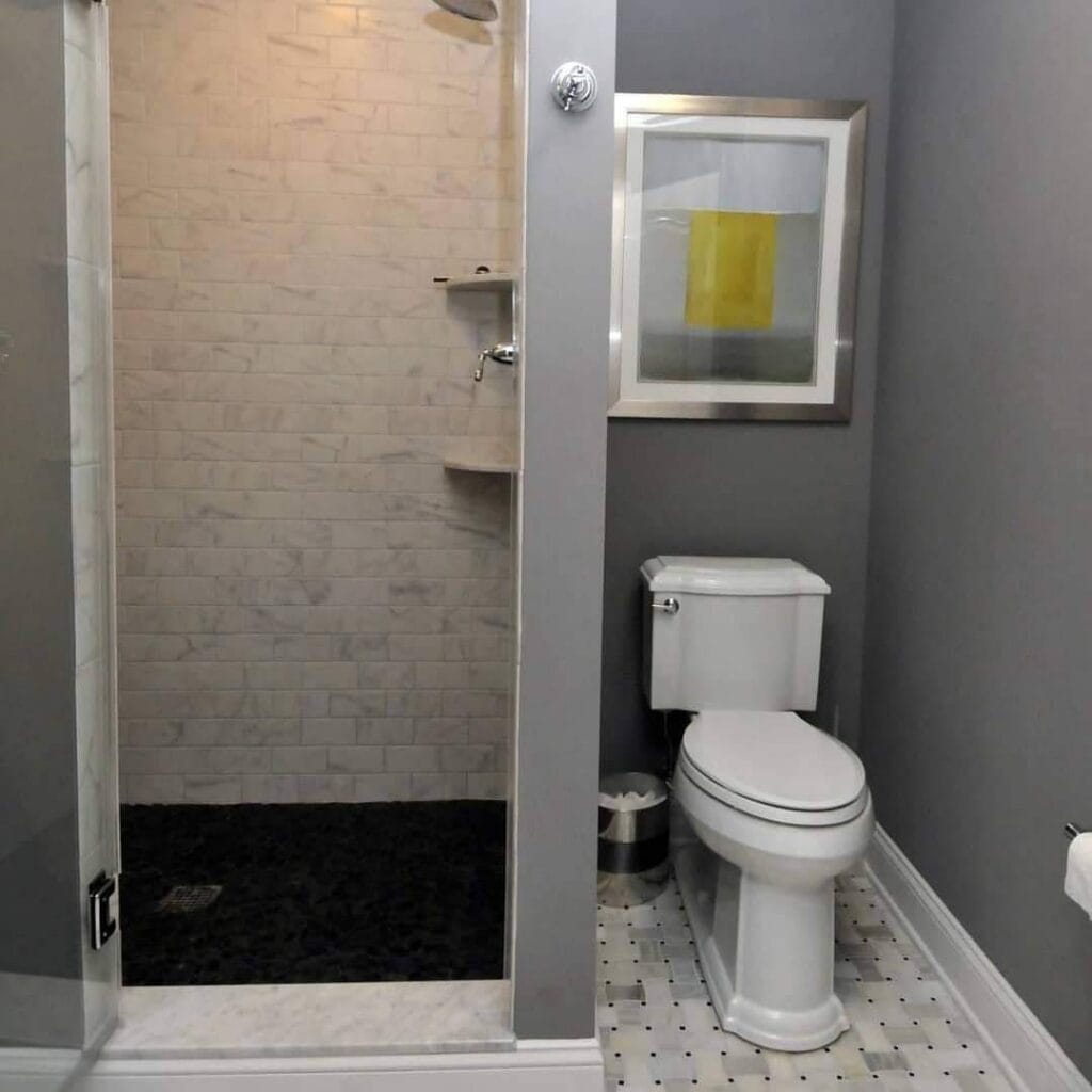 a modern bathroom with gray walls, a glass shower with white subway tiles, a white toilet, framed abstract art above the toilet, and a tiled floor with a small trash can beside the toilet.