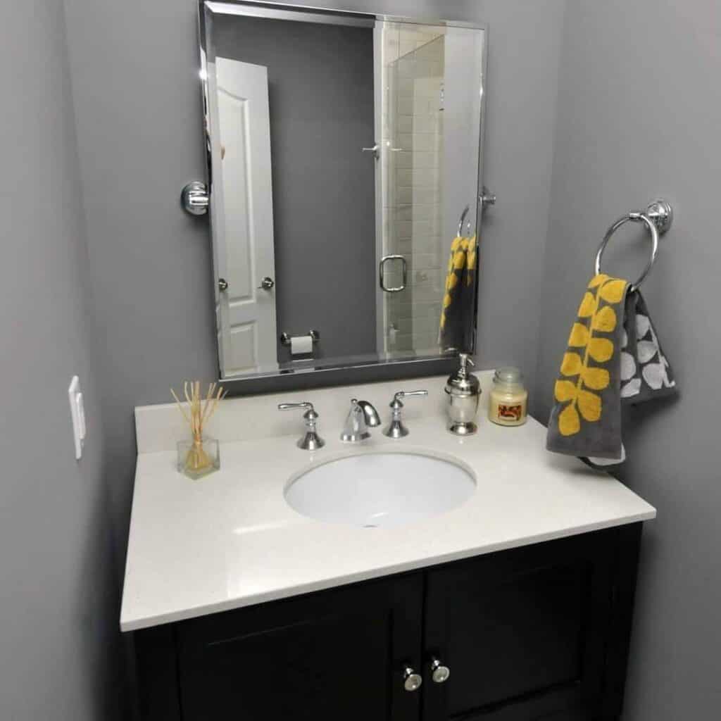 a modern bathroom sink with a white countertop, silver faucet, mirror, and dark cabinet. a gray towel with yellow leaf patterns hangs on a ring. decorative items and a candle are on the counter. gray walls complete the room.