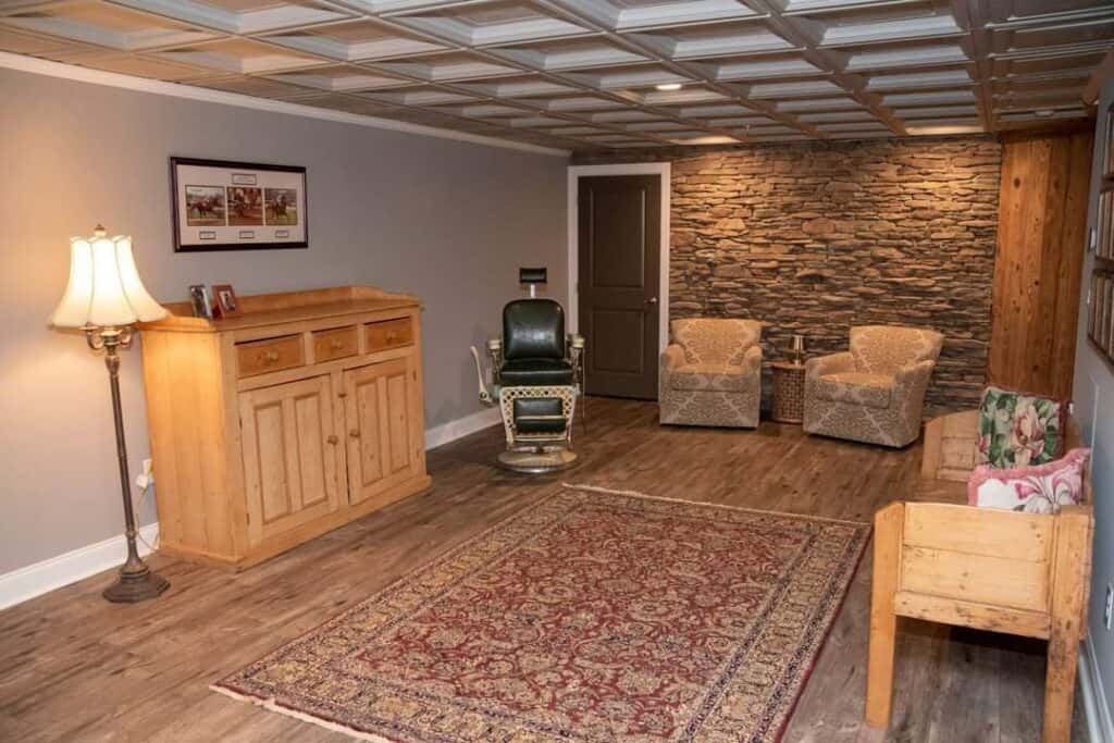 a cozy room with wood flooring, a patterned rug, stone accent wall, two armchairs, a vintage barber chair, wooden sideboard, lamp, and a wooden bench with floral cushions. framed pictures hang on the wall.