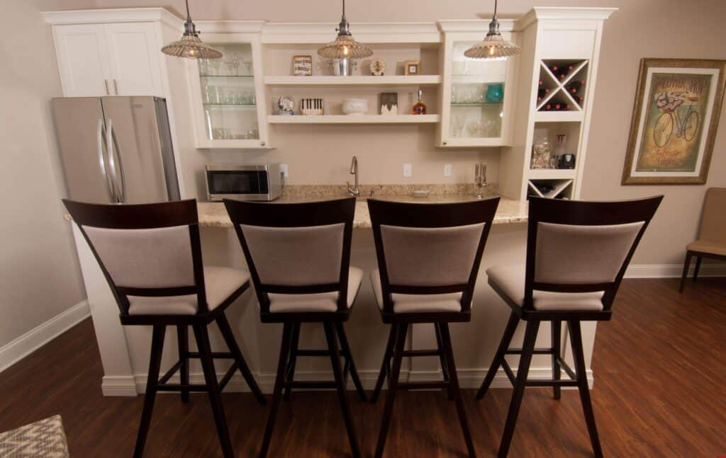 a modern kitchen with four upholstered bar stools lined up at a marble counter, pendant lights above, glass front cabinets, stainless steel refrigerator, and a wall art of a bicycle on the right.