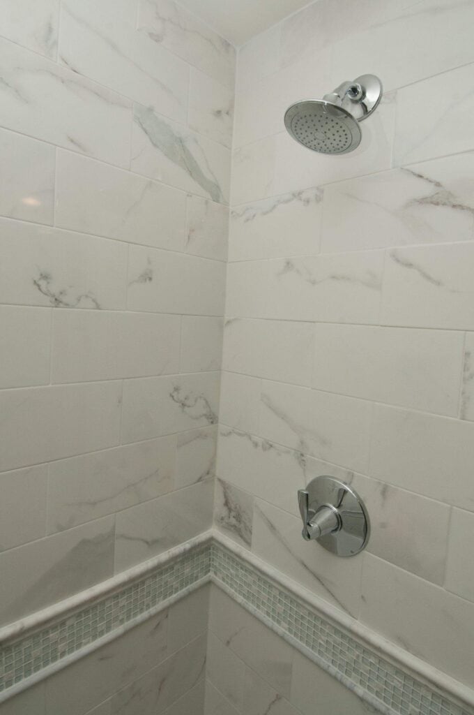 a shower with white marble patterned tiles, a round silver showerhead at the top, and a matching silver control handle below, with a mosaic tile border running horizontally along the wall.