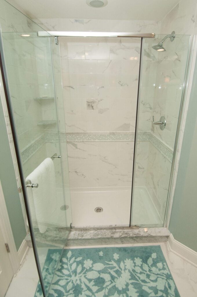 modern shower with glass doors, white marble tile walls, chrome fixtures, and a built in shelf. a white towel hangs on the door. a blue floral bath mat is in front of the entrance.