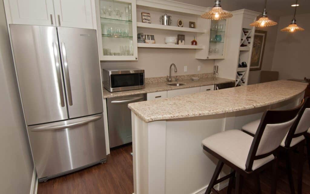 a modern kitchen with a stainless steel refrigerator, microwave, and dishwasher, granite countertops, open shelves with dishes, and a breakfast bar with two cushioned chairs on a wooden floor.
