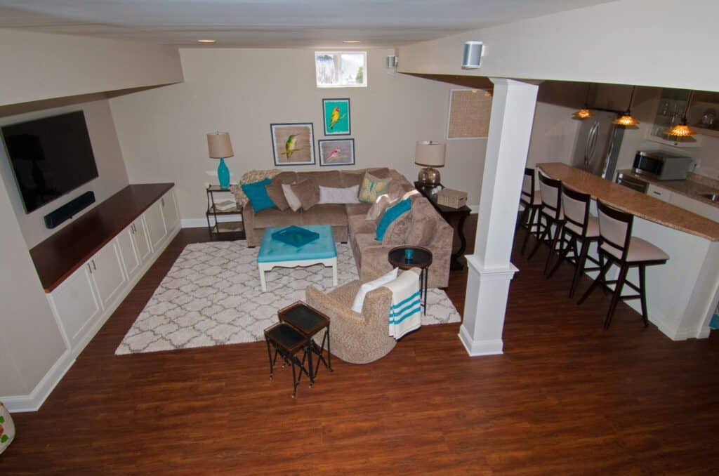 a cozy basement living area with a sectional sofa, turquoise and teal accents, a white coffee table, framed art on the wall, a large tv, bar seating, and a kitchen area with hanging pendant lights and wood flooring.
