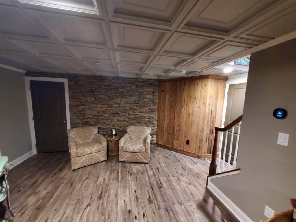 a cozy room with wood patterned flooring, two patterned armchairs around a small round table, a stone accent wall, wood paneled section, and a staircase with white railings. a digital thermostat is on the wall.