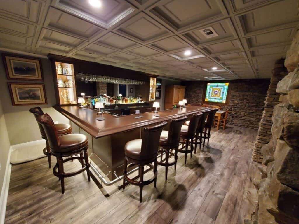 a home bar with a wooden counter, high leather chairs, shelves of glassware and bottles, stone accent walls, framed art, and a stained glass window. there is a small dining table in the corner on wood flooring.
