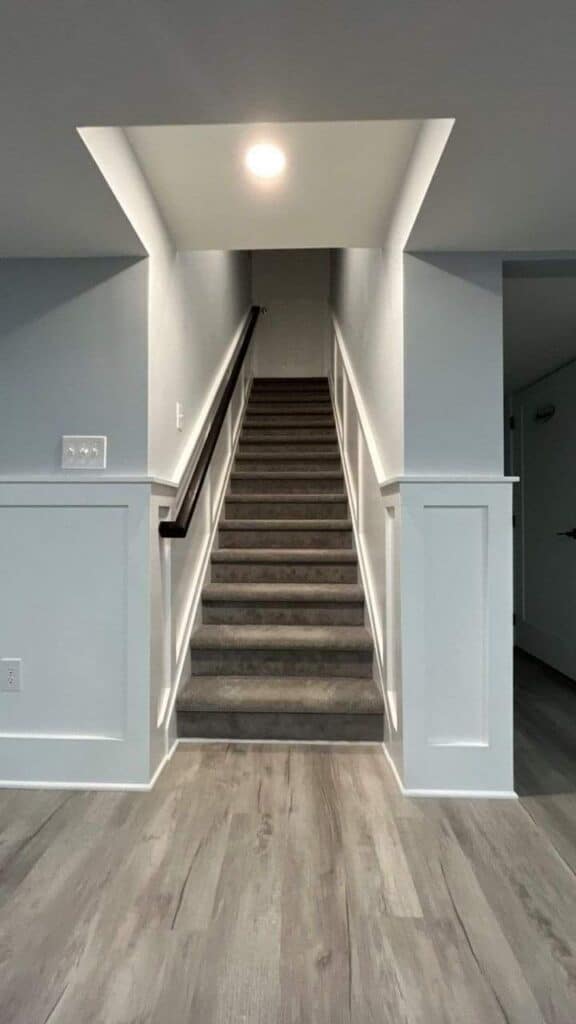 a carpeted staircase with a dark handrail leads upward from a basement with light gray walls, white wainscoting, and light wood style flooring. a ceiling light illuminates the top of the stairs.