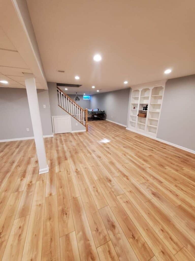 a spacious, finished basement with light wood flooring, recessed lighting, light gray walls, built in white shelves, a staircase with a wooden railing, and a sitting area with a tv in the background.