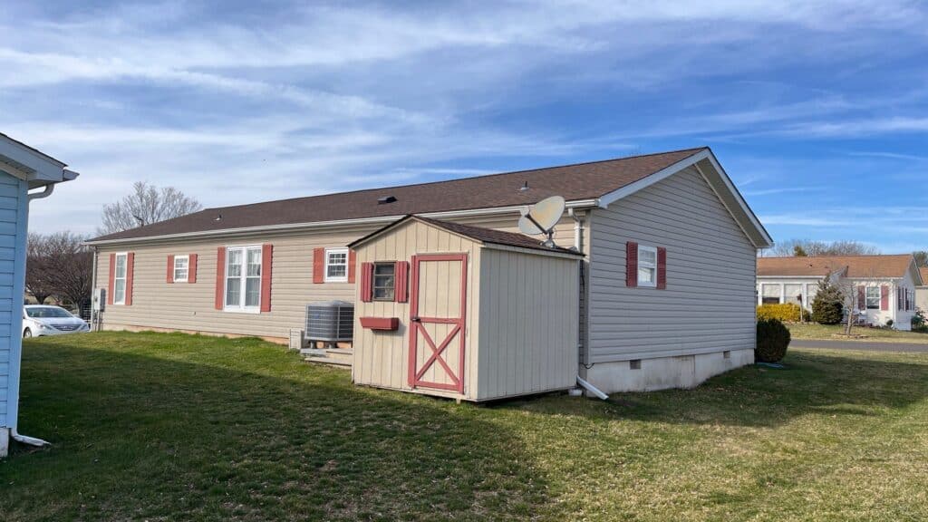 a beige manufactured home with red shutters and a small shed in the yard sits on green grass under a blue sky with wispy clouds. a satellite dish is mounted on the shed, and there are neighboring houses nearby.