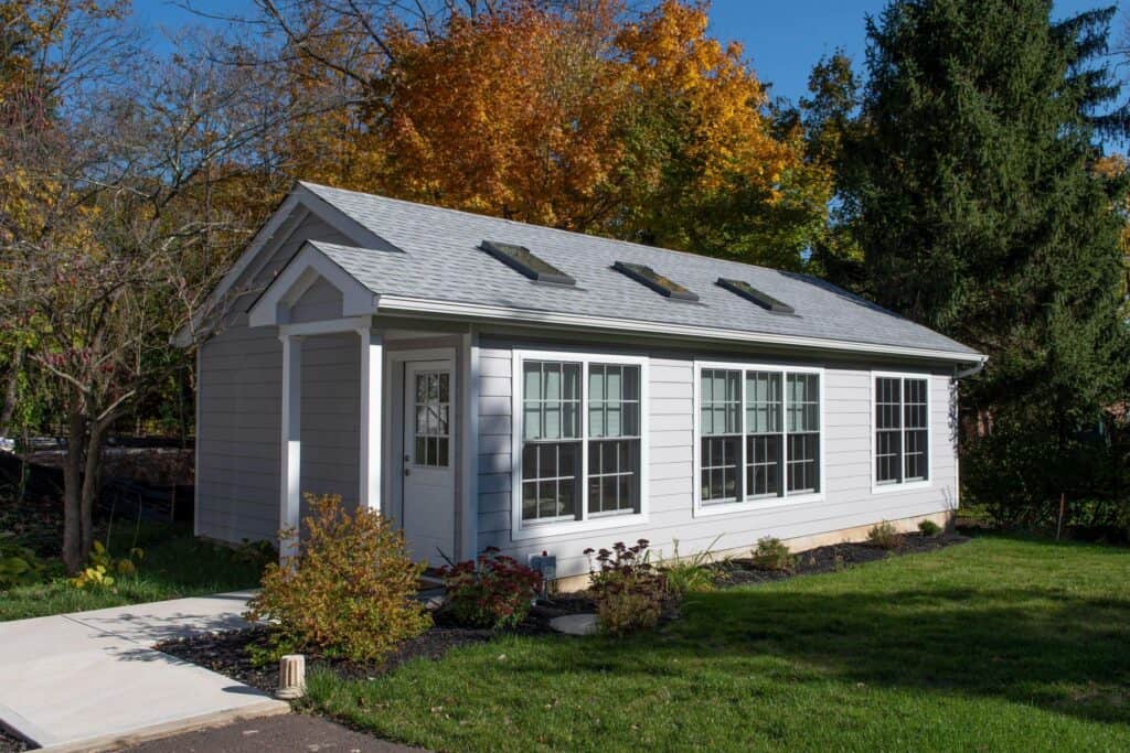 a small, white house with large front windows and three skylights on the roof sits in a grassy yard with shrubs. tall trees with autumn foliage are in the background.
