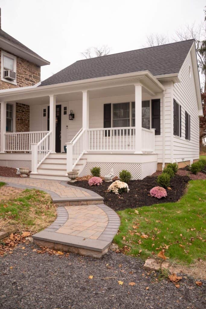 a small white house with a dark roof, black shutters, and a front porch with railings. there are steps leading to a walkway, a lawn, and flower beds with mulch and pink and white flowers.