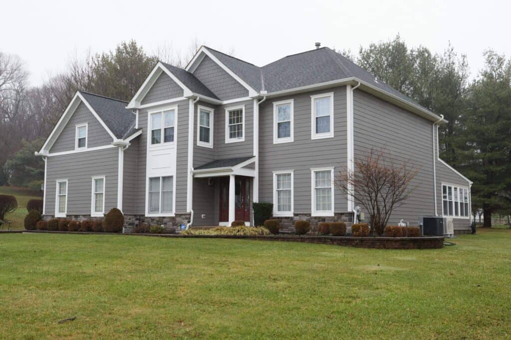 a two story gray house with white trim, multiple gables, and a well maintained lawn. shrubs and trees surround the house, and the sky is overcast, giving a calm and quiet atmosphere.