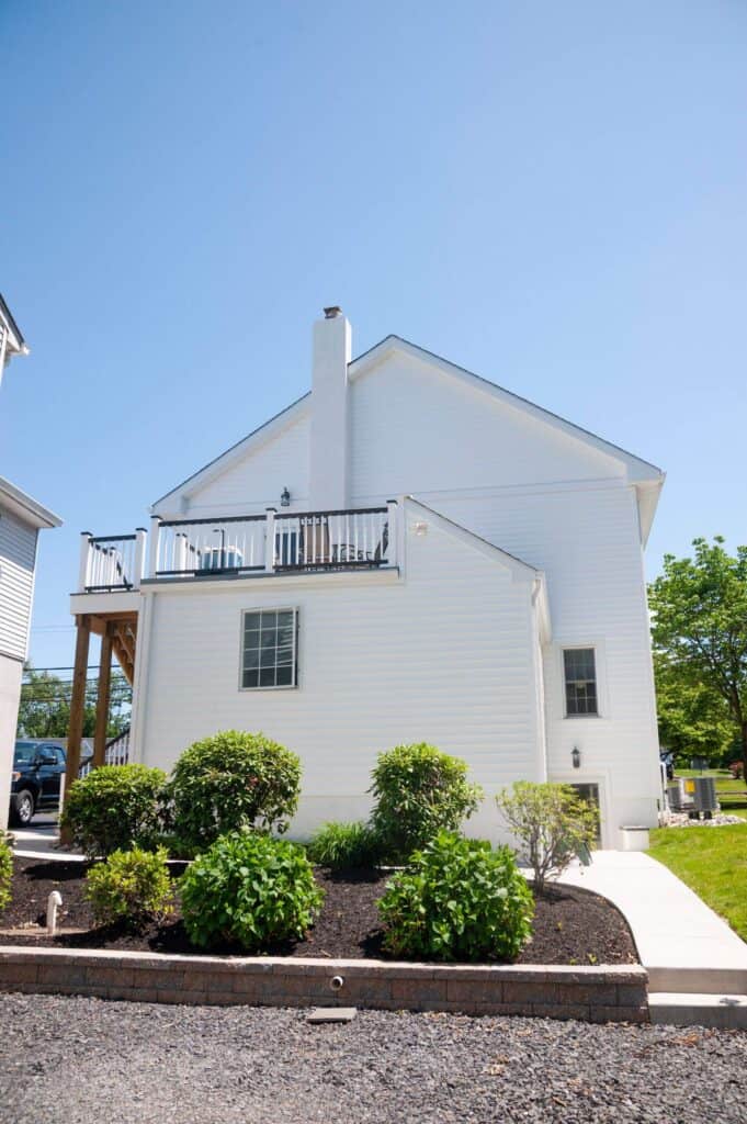 a two story white house with a balcony, surrounded by green bushes and a landscaped garden, under a clear blue sky. a concrete pathway runs along the side of the house.