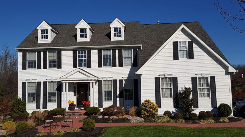 a two story white house with black shutters, dormer windows, and a gray roof, surrounded by neatly trimmed bushes, a brick pathway, and a well maintained green lawn under a clear blue sky.