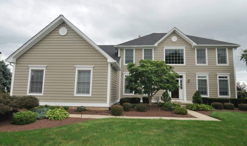 a two story beige house with white trim, multiple windows, and a dark gray roof, surrounded by trimmed bushes, small trees, and a green lawn under a cloudy sky.