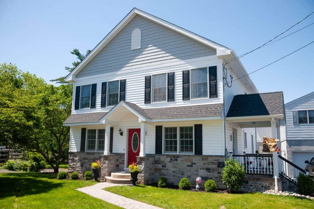 a two story white house with black shutters, a red front door, stone accents, covered porch, and a manicured lawn with potted plants and shrubs on a sunny day.