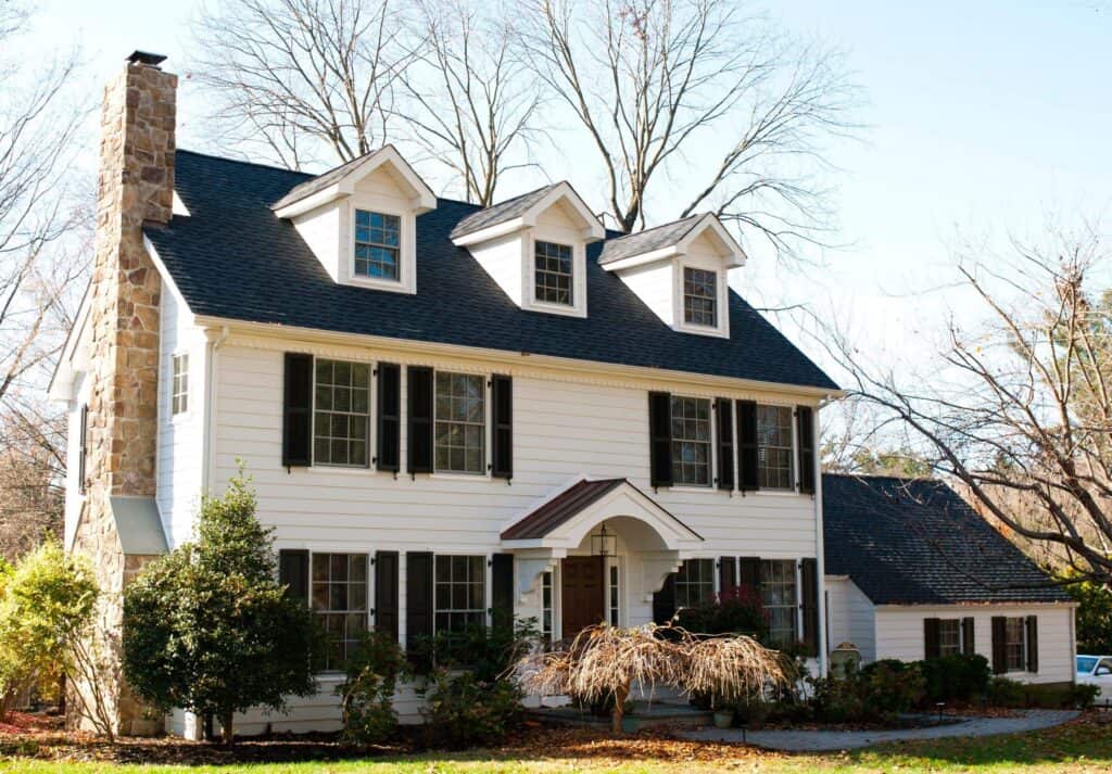 a two story white house with black shutters, three dormer windows, a stone chimney, and a small covered front porch, surrounded by bushes and leafless trees on a sunny day.