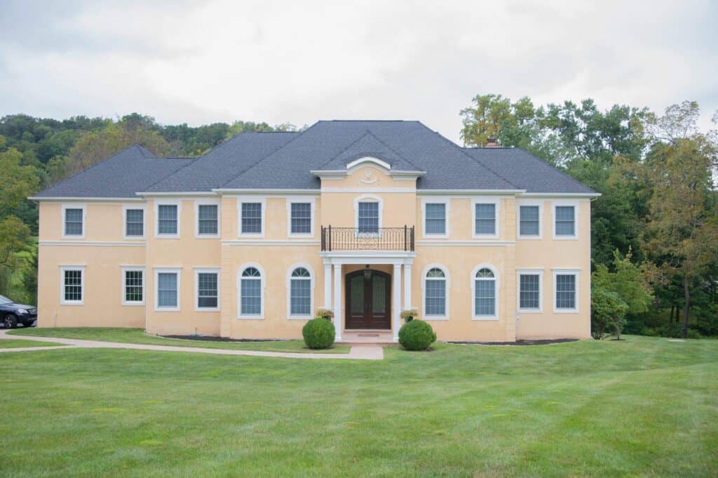 a large, two story yellow house with white trim, multiple windows, a central balcony above the main entrance, and neatly trimmed bushes in front, set on a green lawn with trees in the background.
