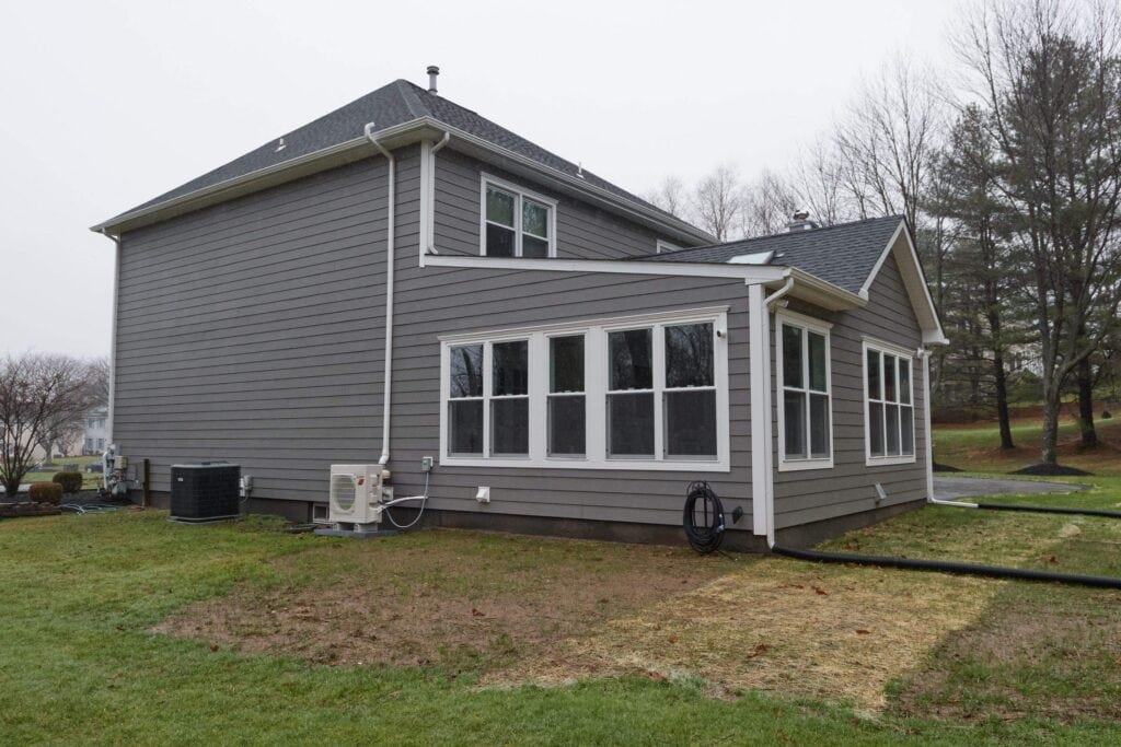 a gray two story house with white trimmed windows sits on a grassy lawn. the back of the house features a sunroom with large windows. trees and other homes are visible in the background on an overcast day.