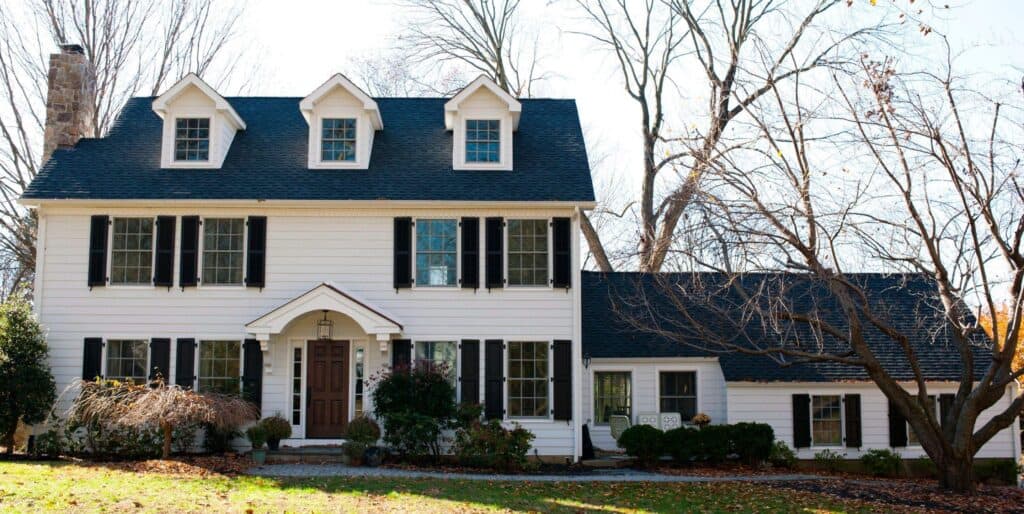 a two story white house with black shutters, three dormer windows on the roof, a central brown front door, and a single story extension on the right, surrounded by trees and shrubs on a sunny day.