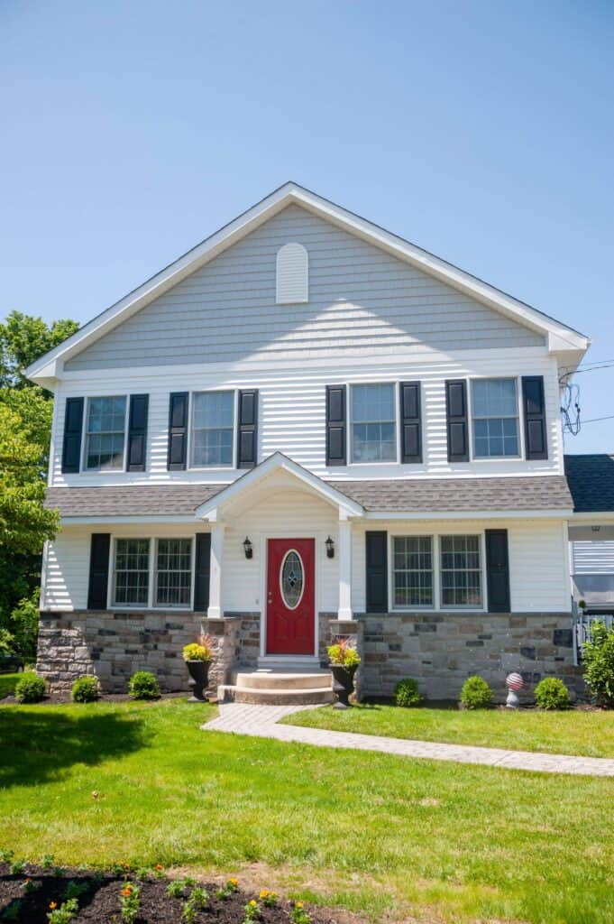 a two story suburban house with white siding, gray stone accents, black shutters, and a red front door. the house has a well kept green lawn, small shrubs, and a clear, sunny sky in the background.
