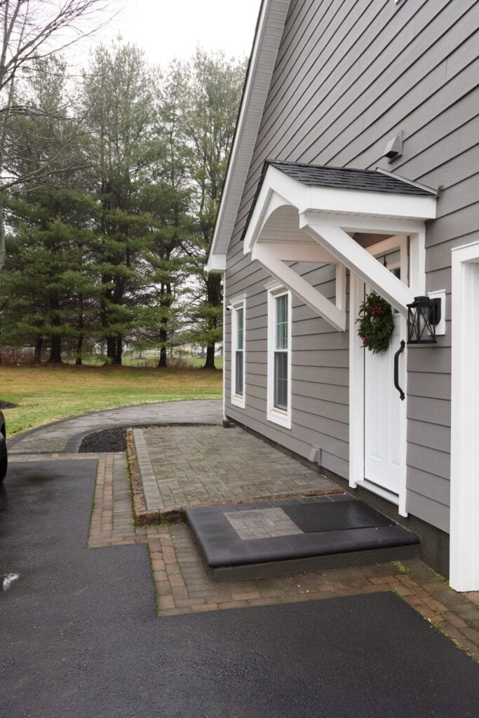 gray house with white trim and a white door decorated with a wreath. the entrance has a small overhang, brick and stone walkway, and is surrounded by trees and a paved driveway.
