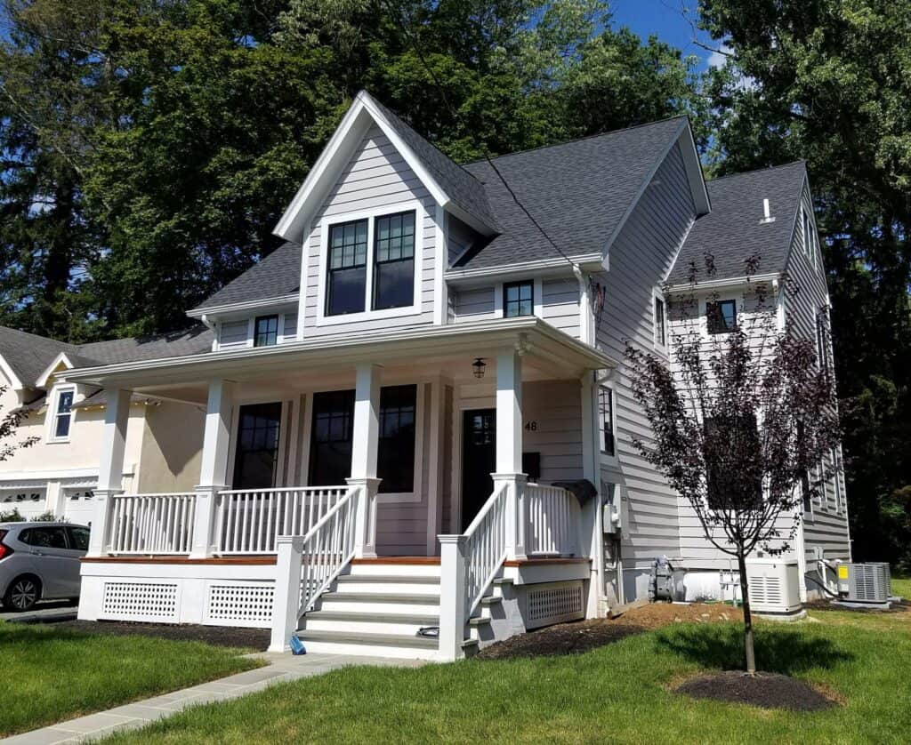 a two story gray house with white trim, a covered front porch, and steps leading up to the entrance, surrounded by green grass and trees, on a sunny day.