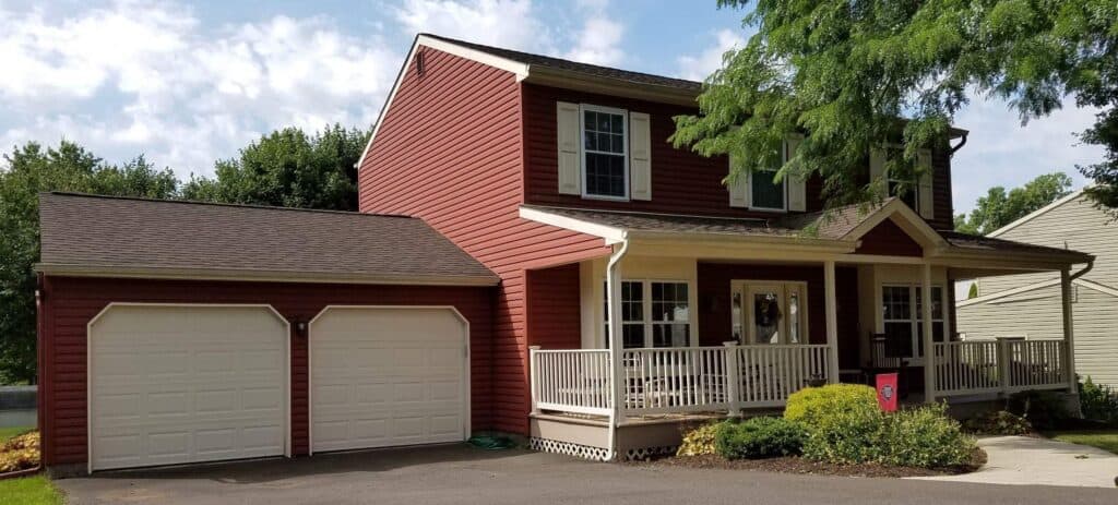 a two story red house with white trim, a covered front porch, and attached two car garage. there are shrubs and a tree in the front yard, and the sky is partly cloudy.