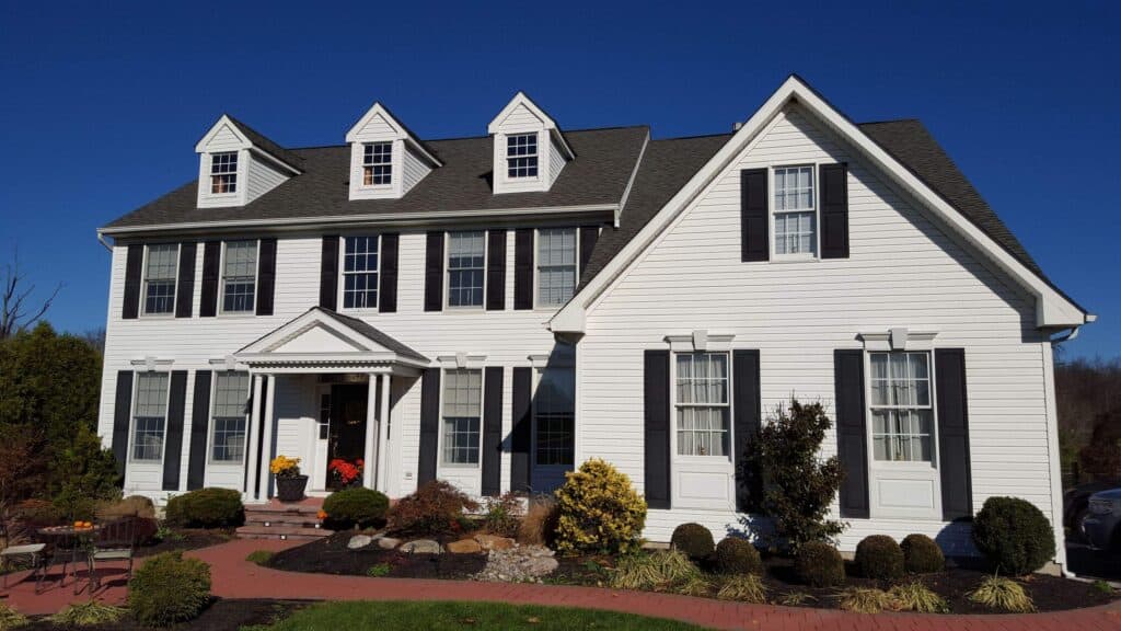a large white two story house with black shutters, dormer windows, and a covered entryway. the front yard has shrubs, small trees, and a brick walkway, all under a clear blue sky.